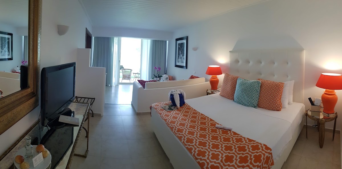 Modern resort bedroom with white bedding, colorful accent pillows, orange lamps, TV, and a balcony overlooking the grounds at Grecotel Creta Palace.