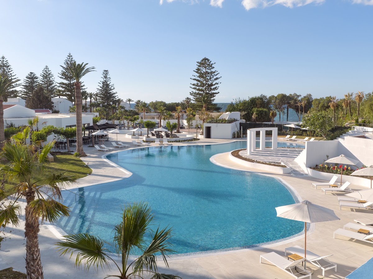 Curved turquoise pool with white loungers and umbrellas surrounded by palm trees and white resort buildings at Grecotel Creta Palace.