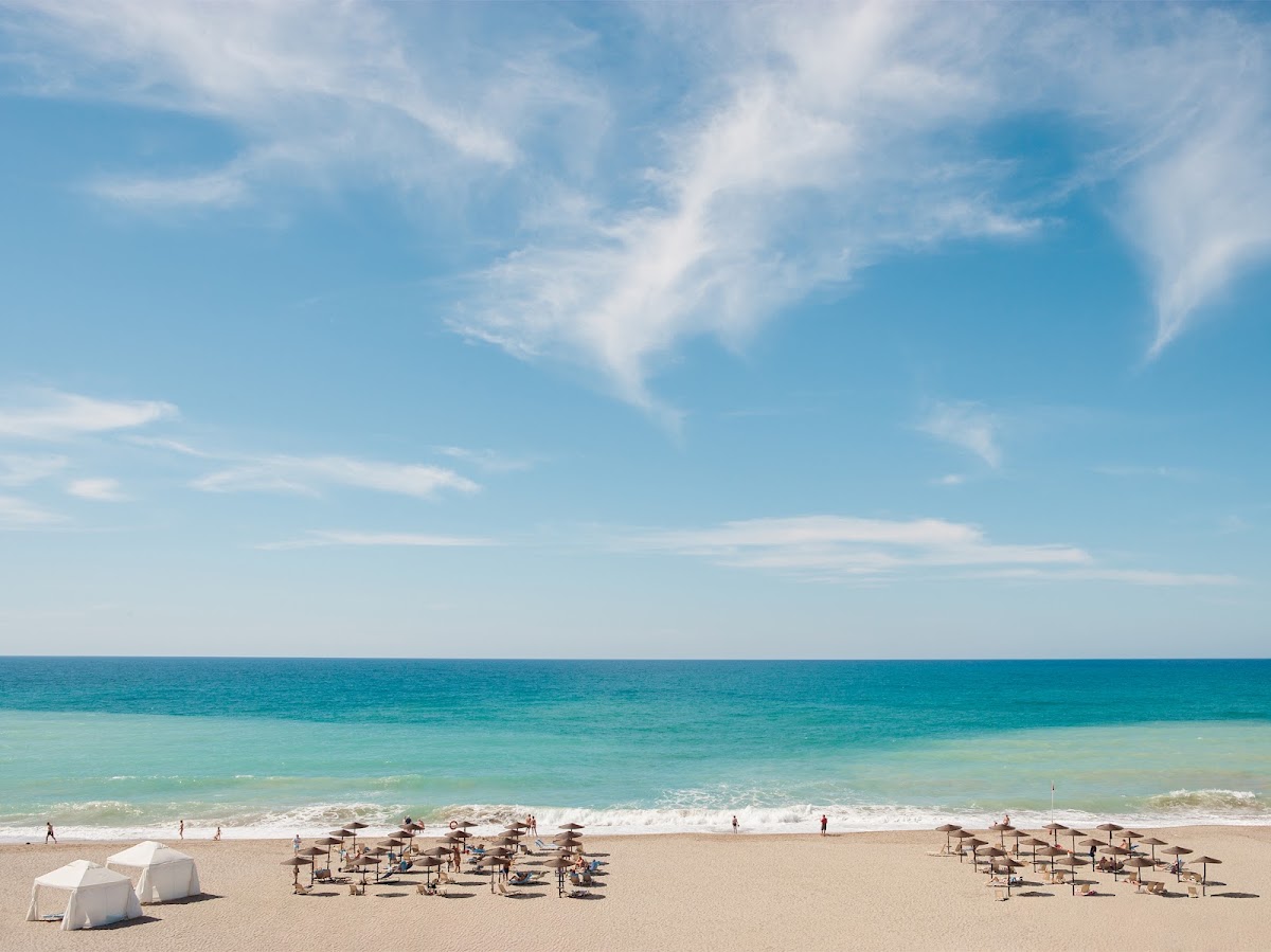 Beach at Grecotel Creta Palace with rows of thatched umbrellas, white loungers, turquoise water, and swimmers under a partly cloudy sky.