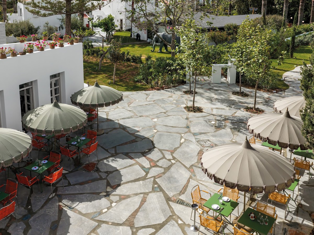 Stone courtyard with red and green dining tables under cream umbrellas, white buildings, and trees at Grecotel Creta Palace.