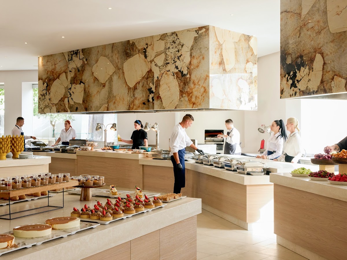 Open kitchen with staff preparing food at multiple stations, featuring marble countertops and a large abstract stone accent wall above.