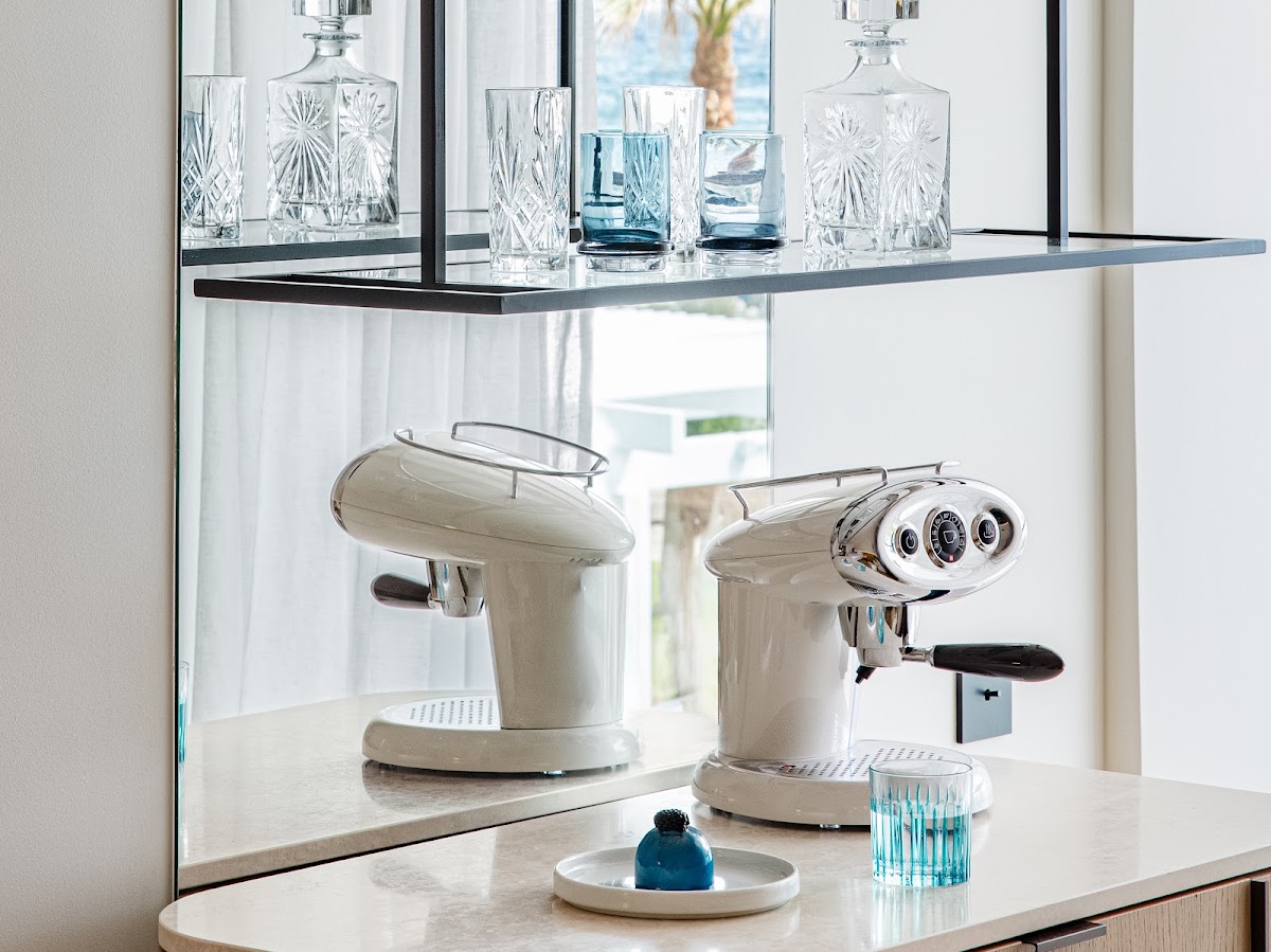 Modern in-room bar setup with cream espresso machines, crystal glassware, and ocean view through window at Grecotel Creta Palace.