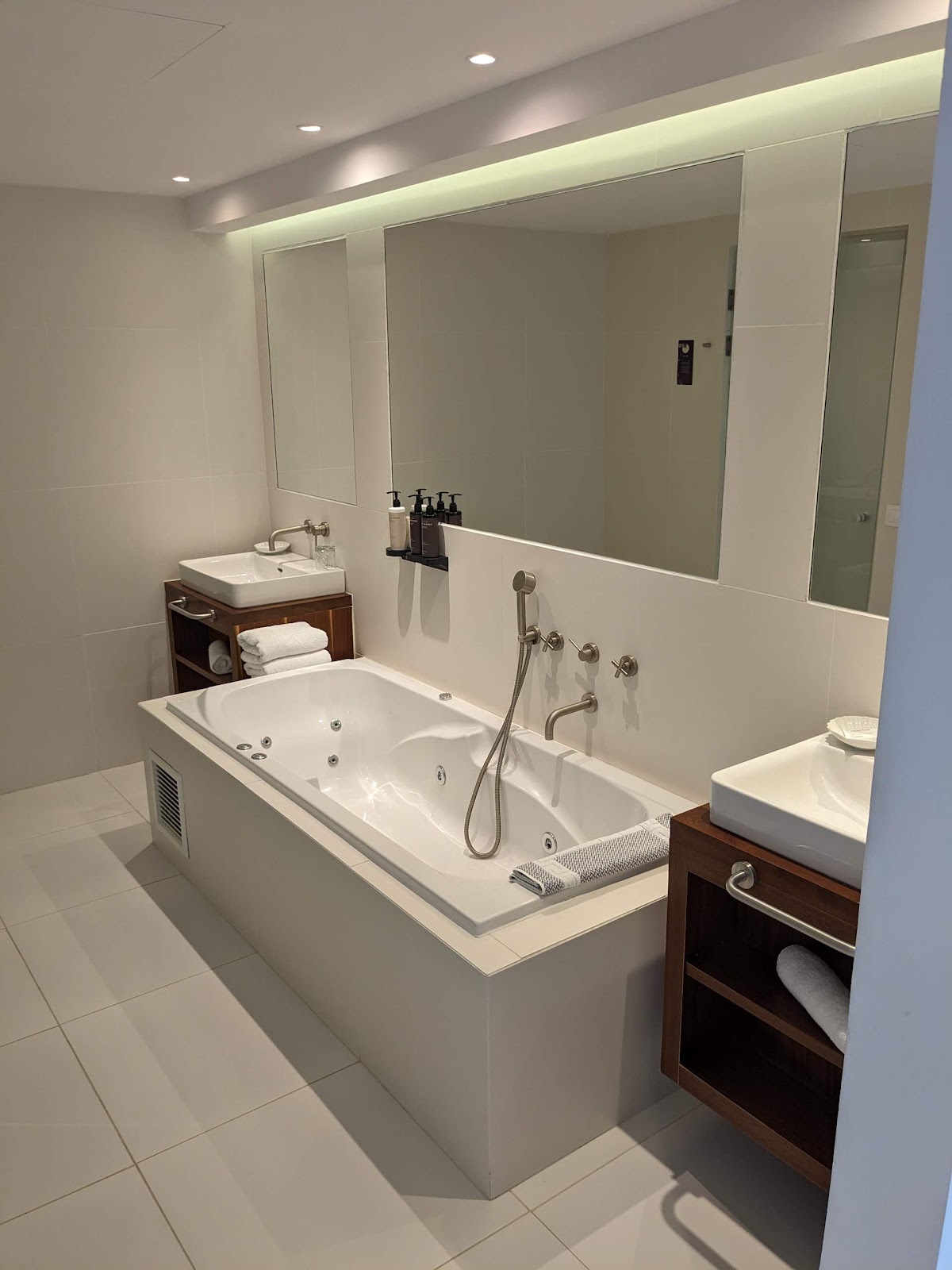 Modern bathroom with white soaking tub, dual vanity sinks, large illuminated mirror, and light tile flooring at Grecotel LUXME White.
