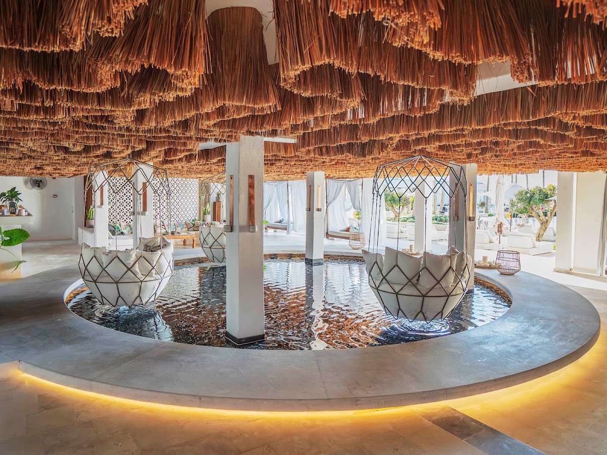 Covered lobby area with hanging copper-toned fringe ceiling, circular water feature with decorative baskets, white columns, and open views to beach beyond.