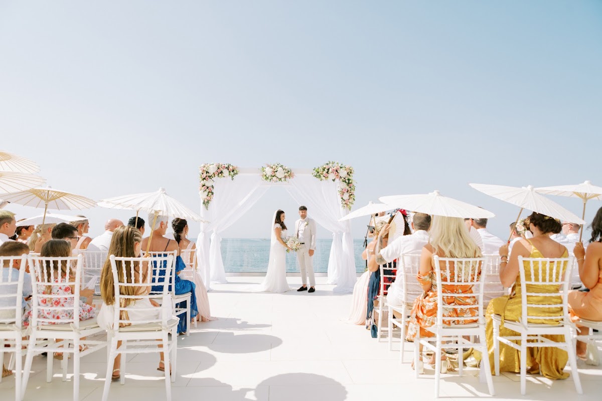 Bride and groom stand at white beachside wedding altar with guests seated under white umbrellas and a clear blue sky.