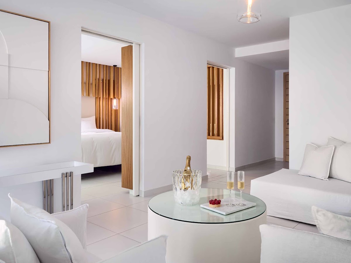 Modern suite living area with white furnishings, glass table with champagne bucket, and wooden-accented bedroom visible through open doorway.