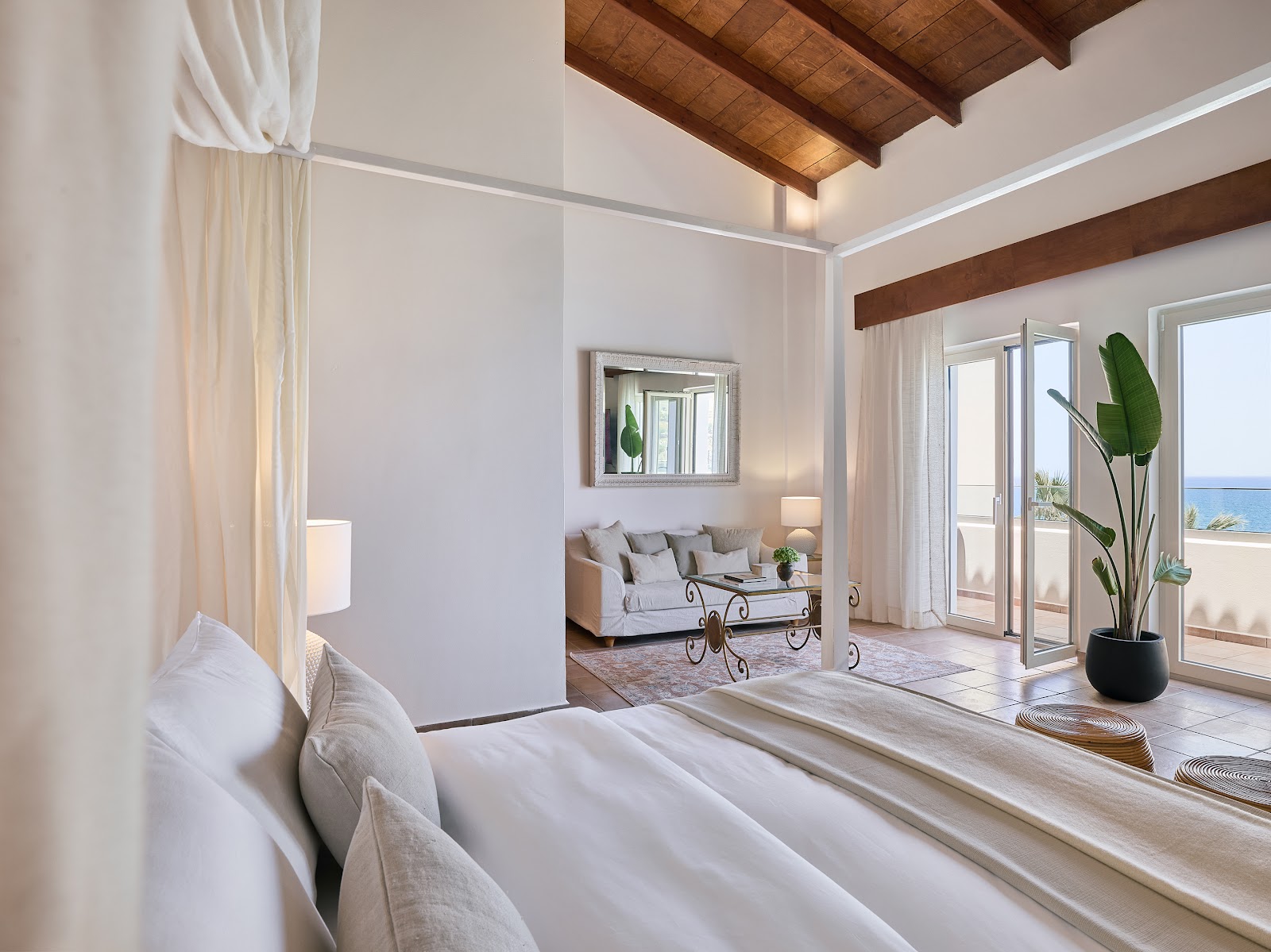 Bright bedroom with white linens, wooden beams, and glass doors opening to a sea view terrace at Grecotel Marine Palace & Aqua Park.