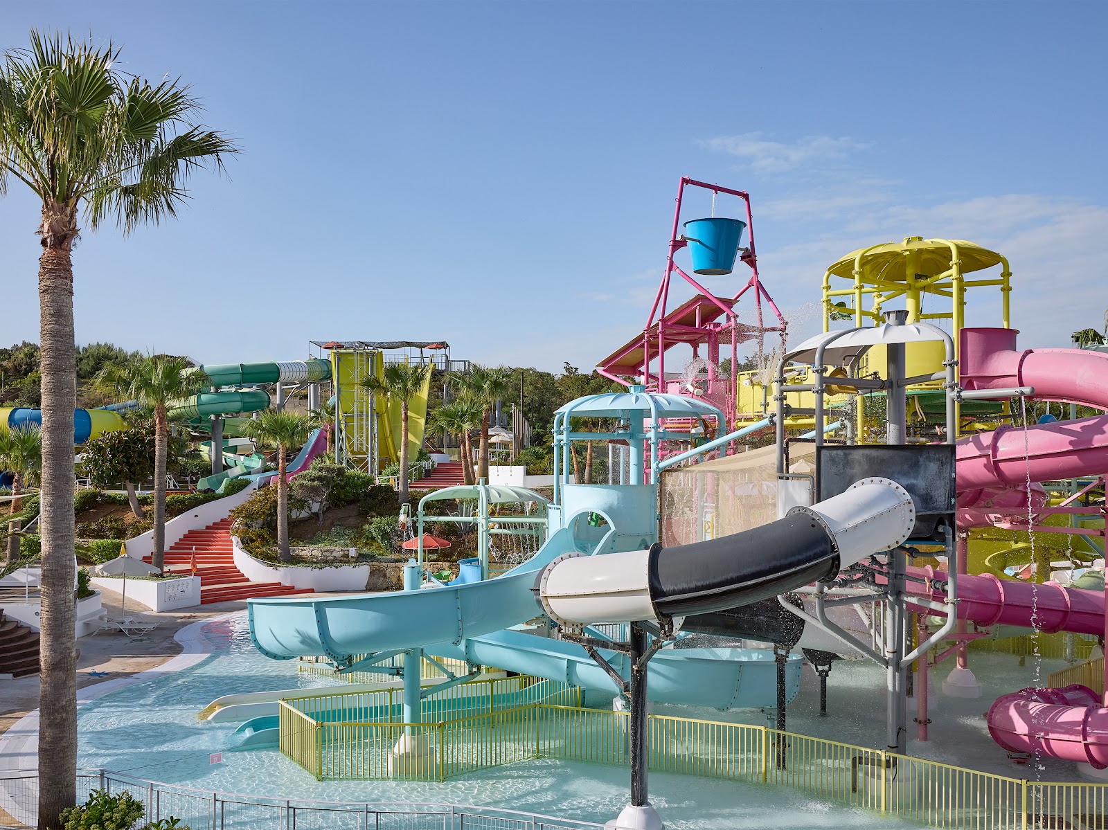 Colorful water slides and tower structures at Grecotel Marine Palace & Aqua Park, with shallow pools, palm trees, and clear sky.