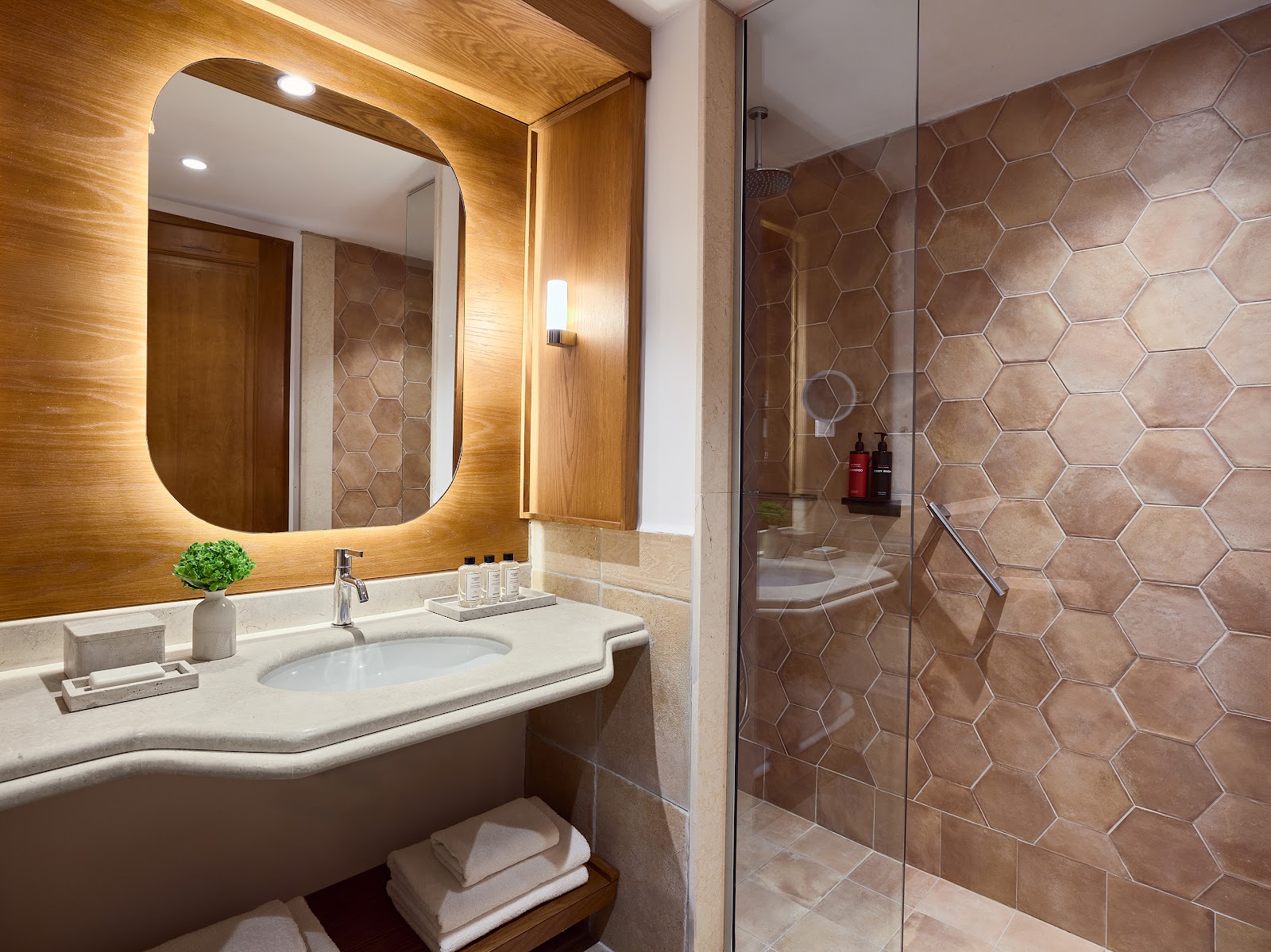 Bathroom with illuminated wooden-framed mirror, white curved sink, hexagonal tile shower enclosure, and folded towels on shelf.