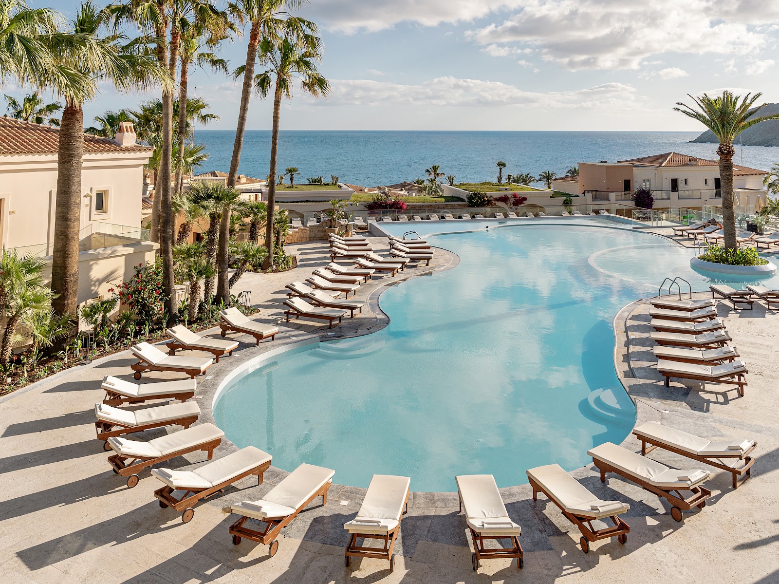 Turquoise pool with island bar surrounded by loungers, palm trees, and beige resort buildings overlooking a rocky coastline and blue sea.