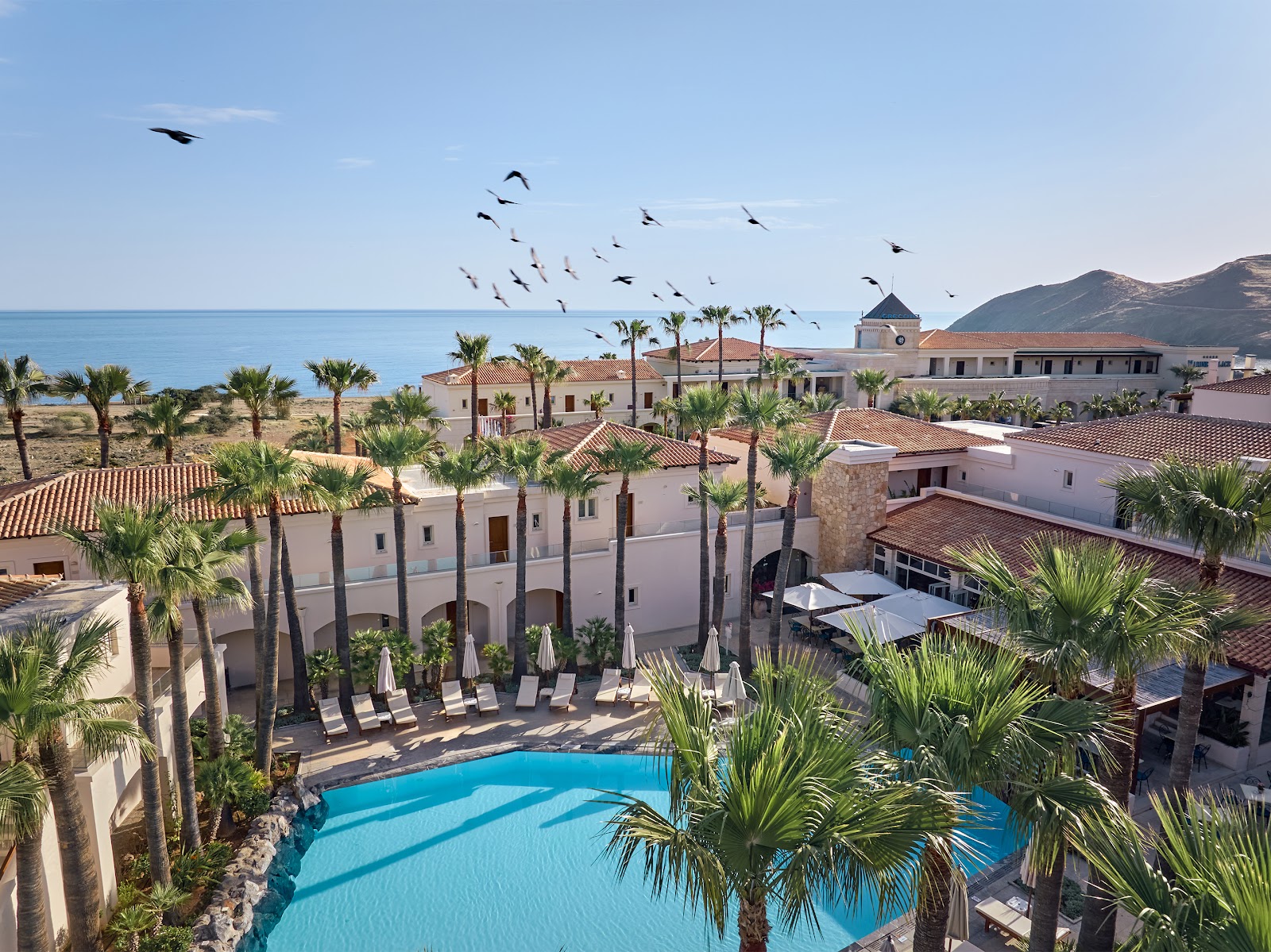 Palm trees frame the white-walled Casa Marina building at Grecotel Marine Palace & Aqua Park, with terracotta-roofed villas visible behind under clear blue sky.