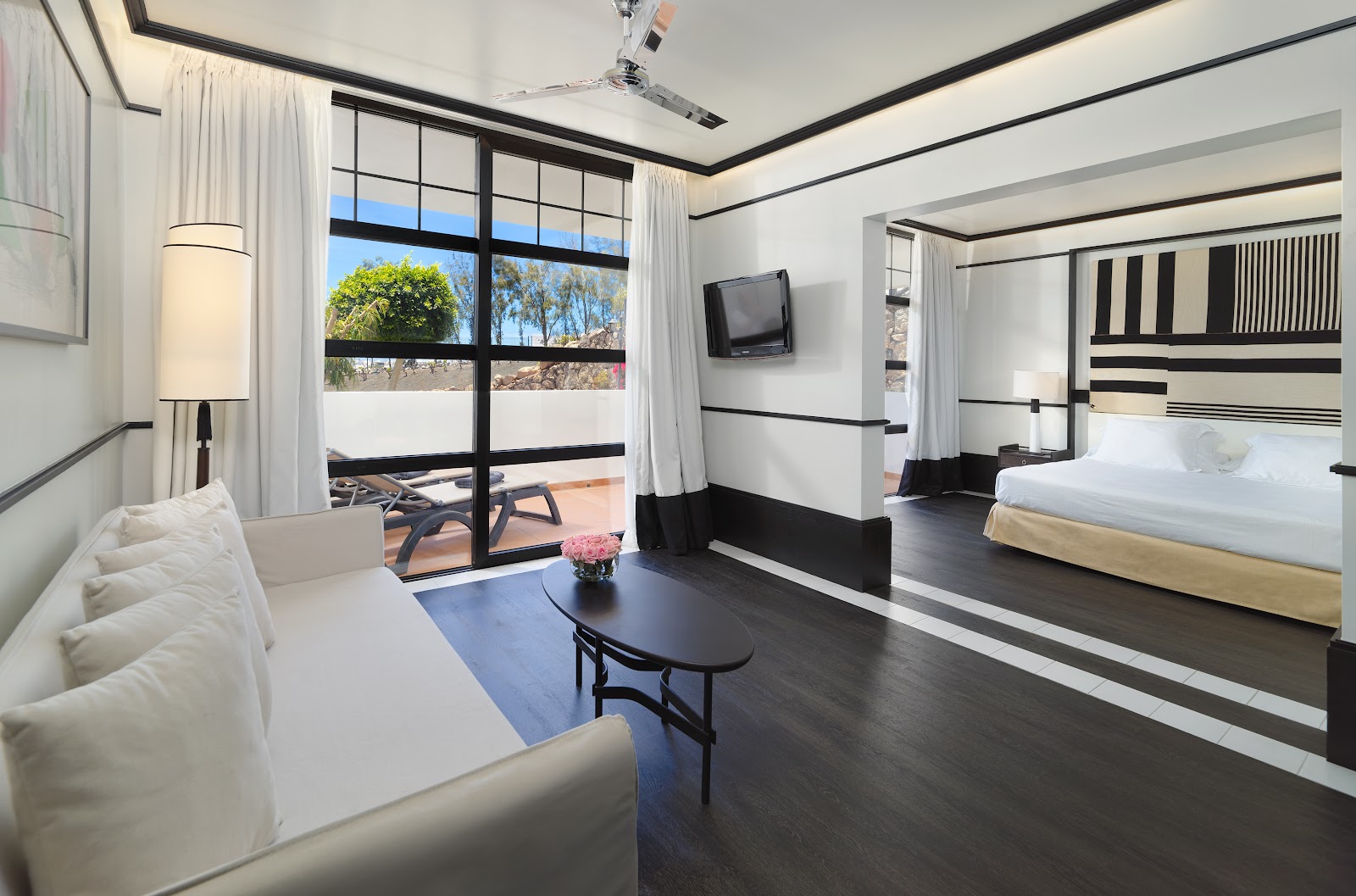 Modern resort suite with white sofa, dark wood floors, and large windows overlooking a terrace with ocean views and landscaping.