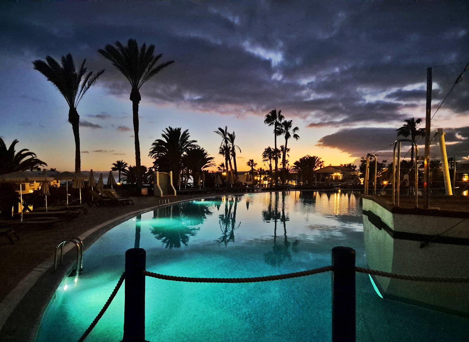 Illuminated pool at dusk with palm trees and resort buildings silhouetted against a colorful twilight sky at H10 Rubicón Horizons Collection.