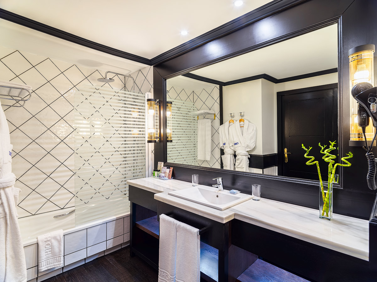 Modern resort bathroom with black-framed mirror, white marble countertop, glass shower enclosure, and green bamboo plant in vase.