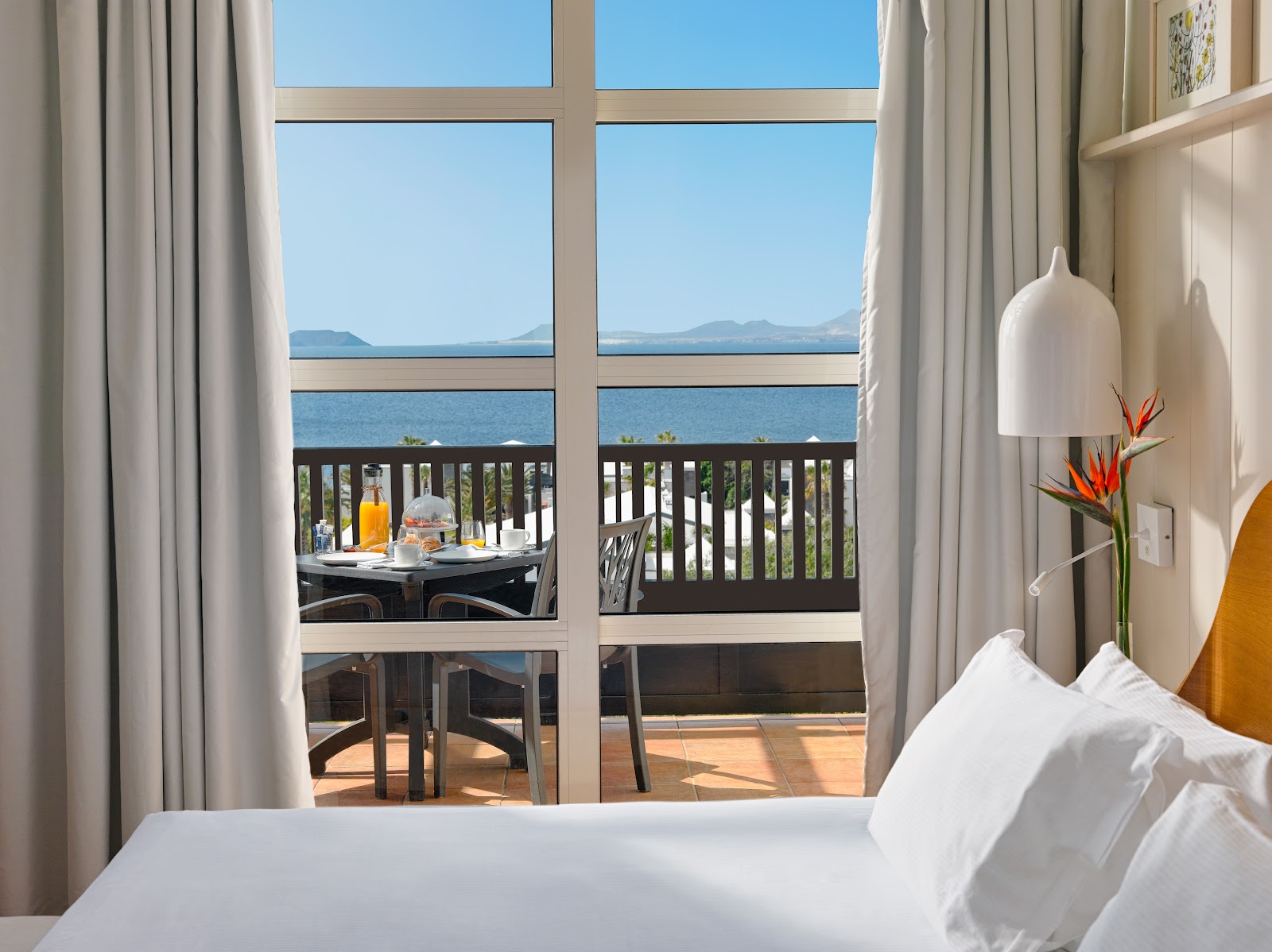 Resort bedroom with ocean view through large windows showing a balcony table, distant mountains, and blue sea under clear sky.