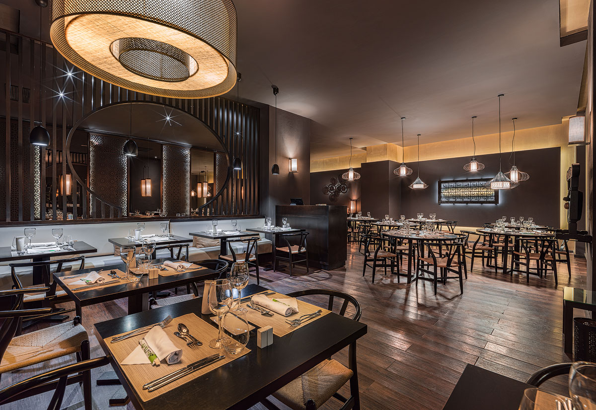 Modern restaurant dining room with dark wood tables set for service, pendant lighting, and a mirrored bar area at H10 Rubicón Horizons Collection.