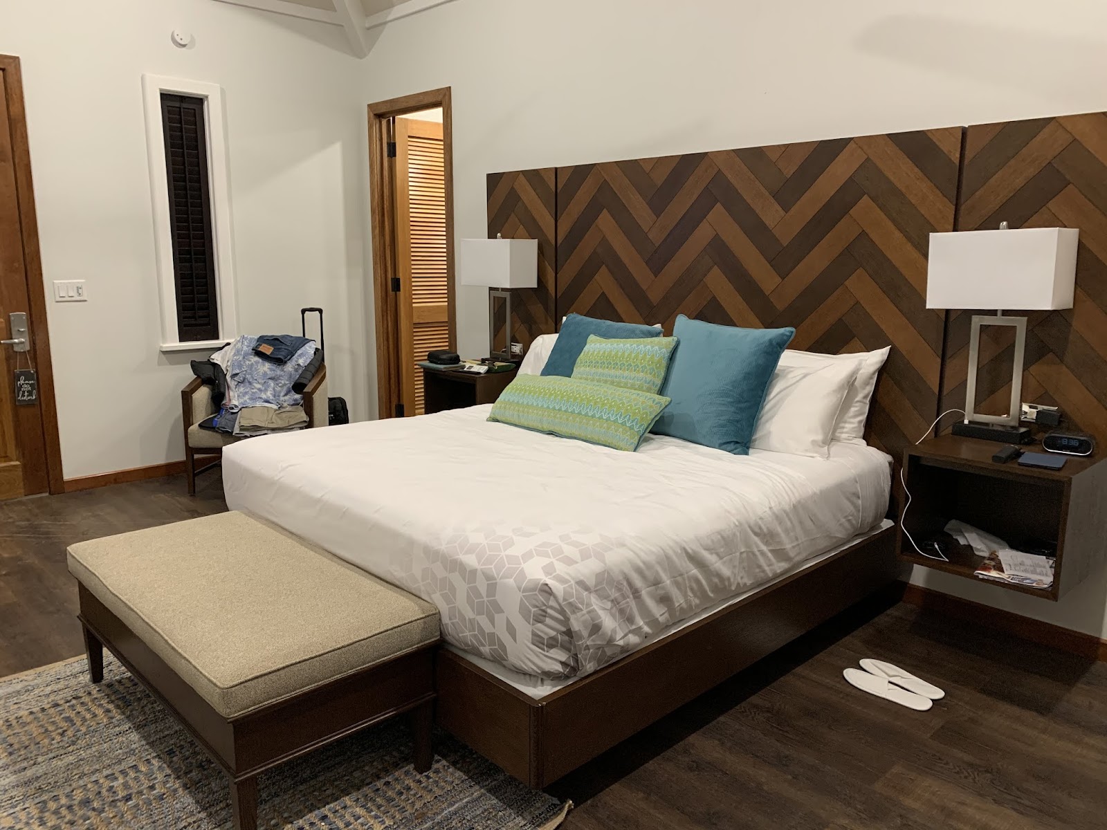 Resort guestroom with queen bed, geometric wood headboard, teal and green pillows, nightstands with lamps, and dark wood furnishings.