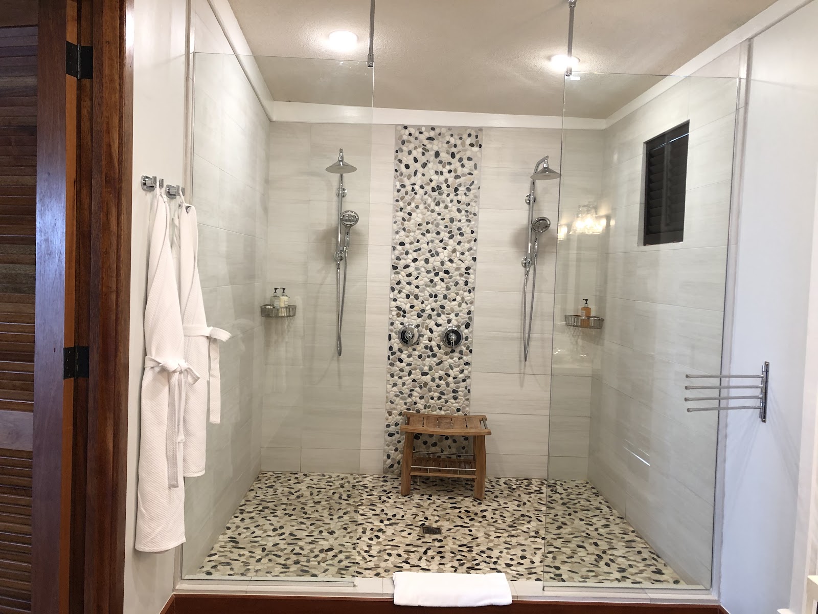 Large walk-in shower with dual showerheads, white tile walls, black and white pebble accent stripe, matching pebble floor, glass enclosure, and white towels on rack.