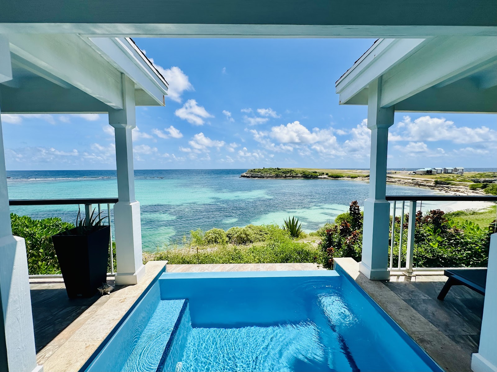 Beachfront cottages at Hammock Cove Antigua line a rocky shoreline overlooking turquoise water under a blue sky with white clouds.