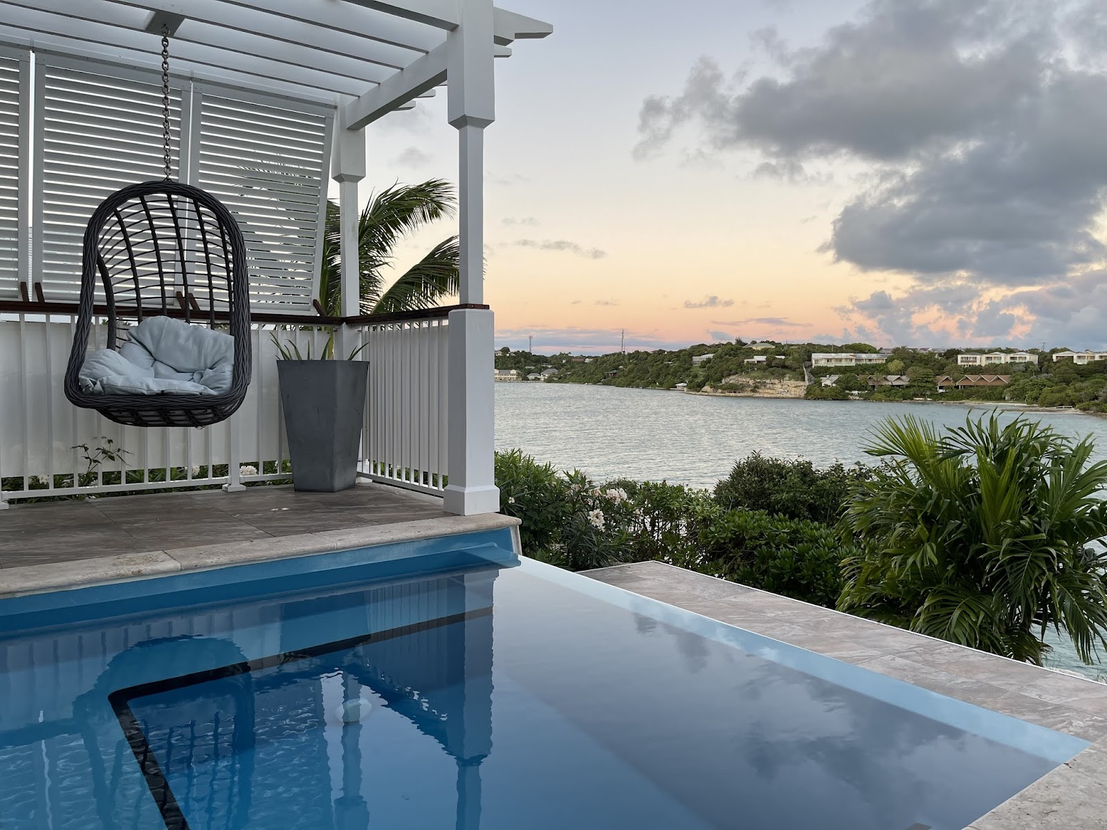 Beach area at dusk with rows of white loungers and beige umbrellas, stone structures, and hillside bungalows at Hammock Cove Antigua.