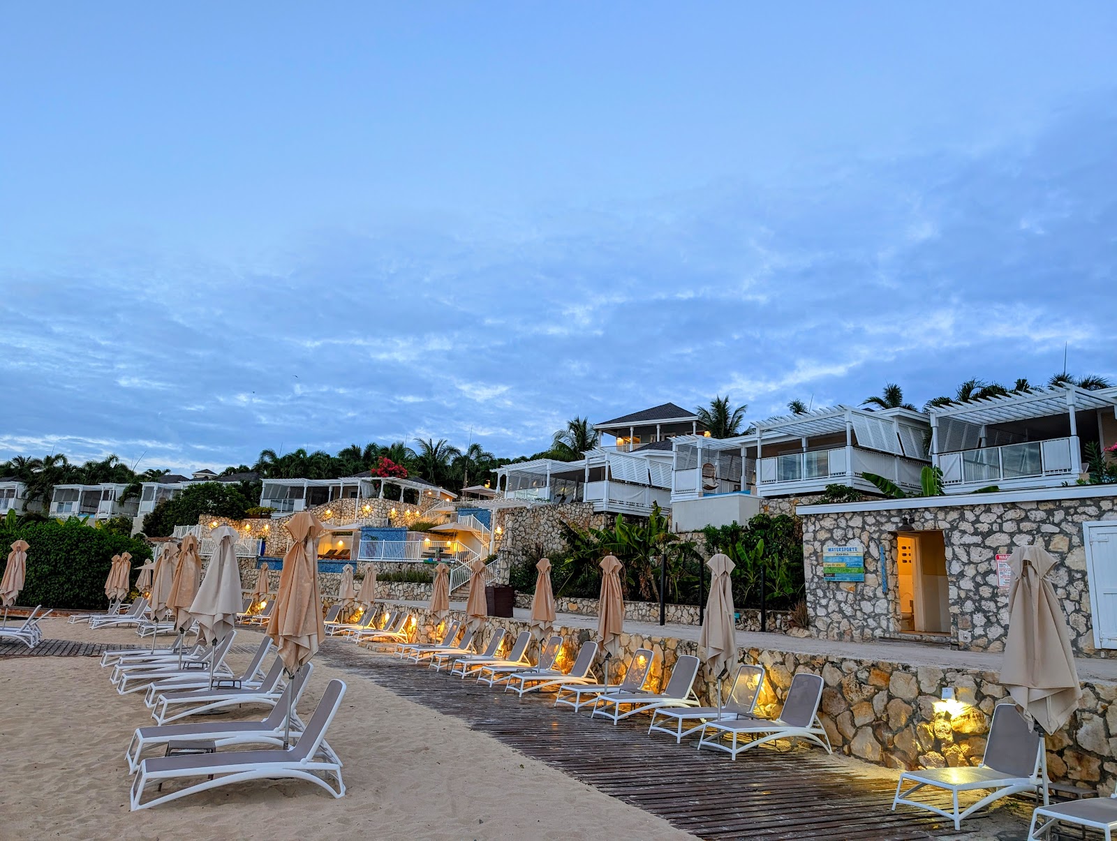 Oceanfront pool deck with blue plunge pool, woven hanging chair, white pergolas, and turquoise Caribbean waters beyond rocky coastline.