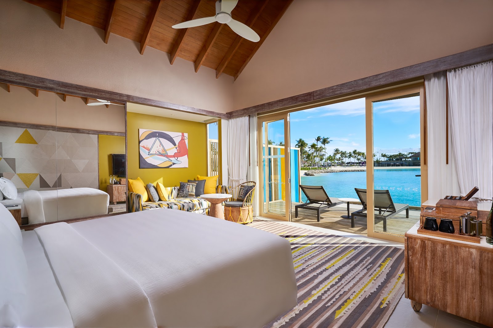 Resort bungalow bedroom with yellow accent wall, wooden beam ceiling, and ocean view through glass doors opening to a private deck with lounge chairs.