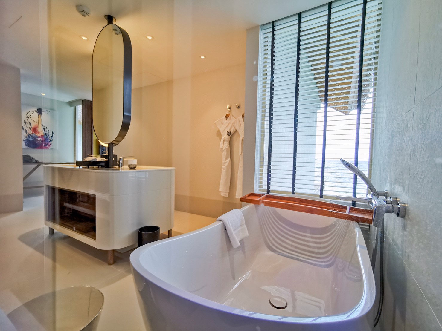 Modern bathroom with curved white soaking tub, oval mirror above vanity with round sink, and louvered windows overlooking ocean at Hard Rock Hotel Maldives.