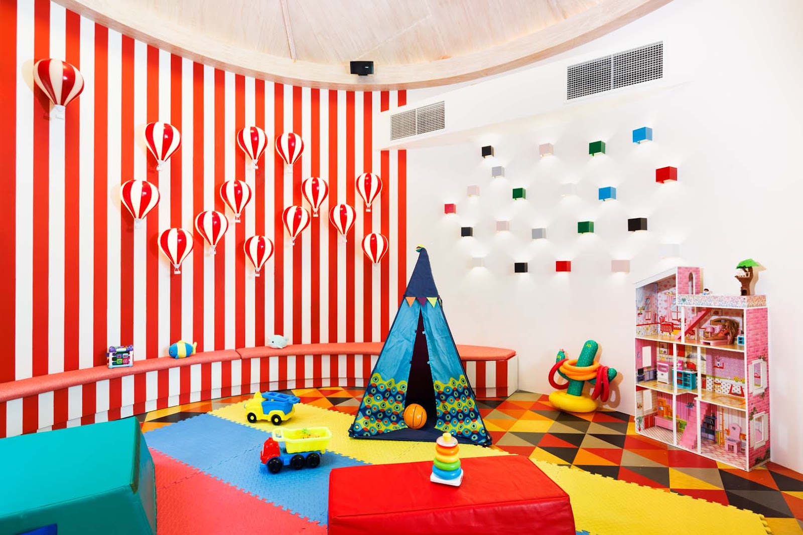 Colorful kids club with red and white striped walls, hanging heart decorations, a blue play tent, toy vehicles, a pink dollhouse, and foam floor mats at Hard Rock Hotel Maldives.