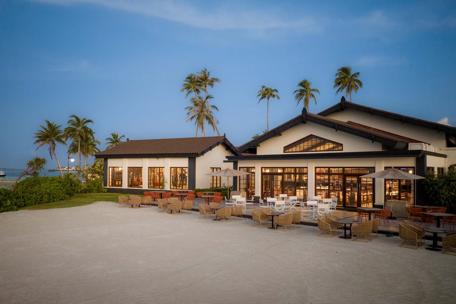 Beachfront dining pavilion at Hard Rock Hotel Maldives with white and dark timber buildings, woven seating, and palm trees at dusk.