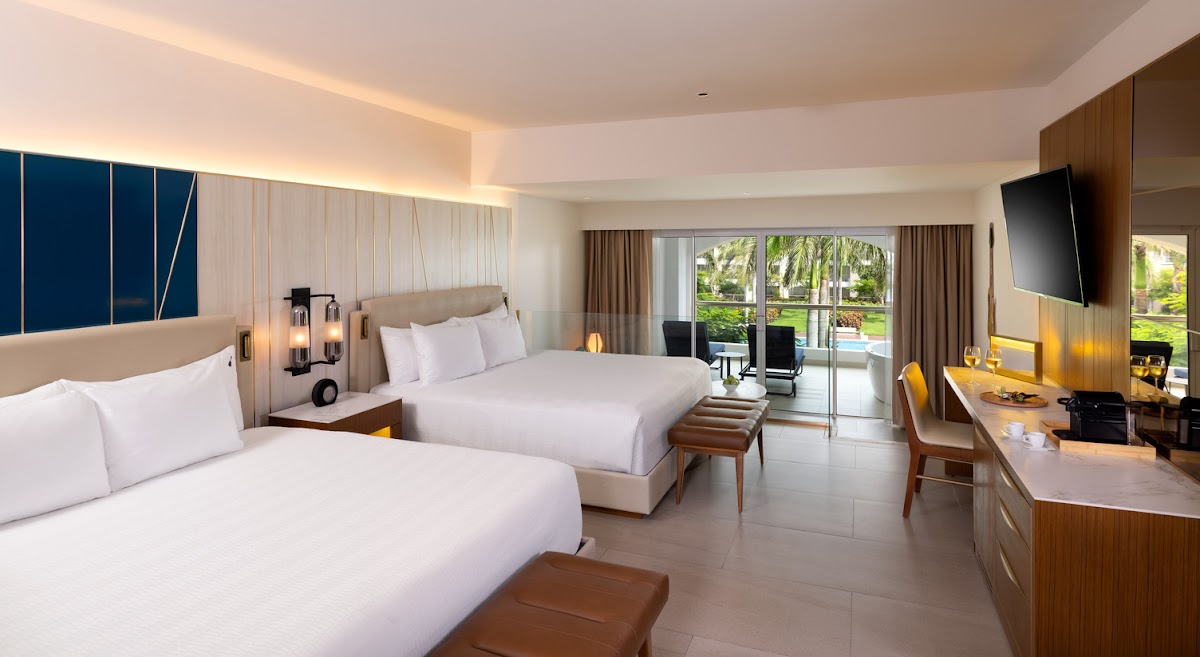 Modern resort room with two queen beds, wood accents, wall-mounted TV, and glass doors opening to pool and tropical grounds.
