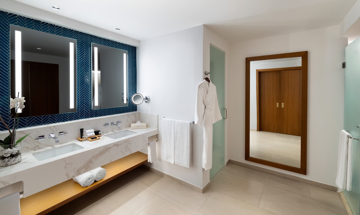 Modern bathroom with dual lit mirrors, marble countertop, white bathtub, and wooden doors at Hard Rock Hotel & Casino Punta Cana.