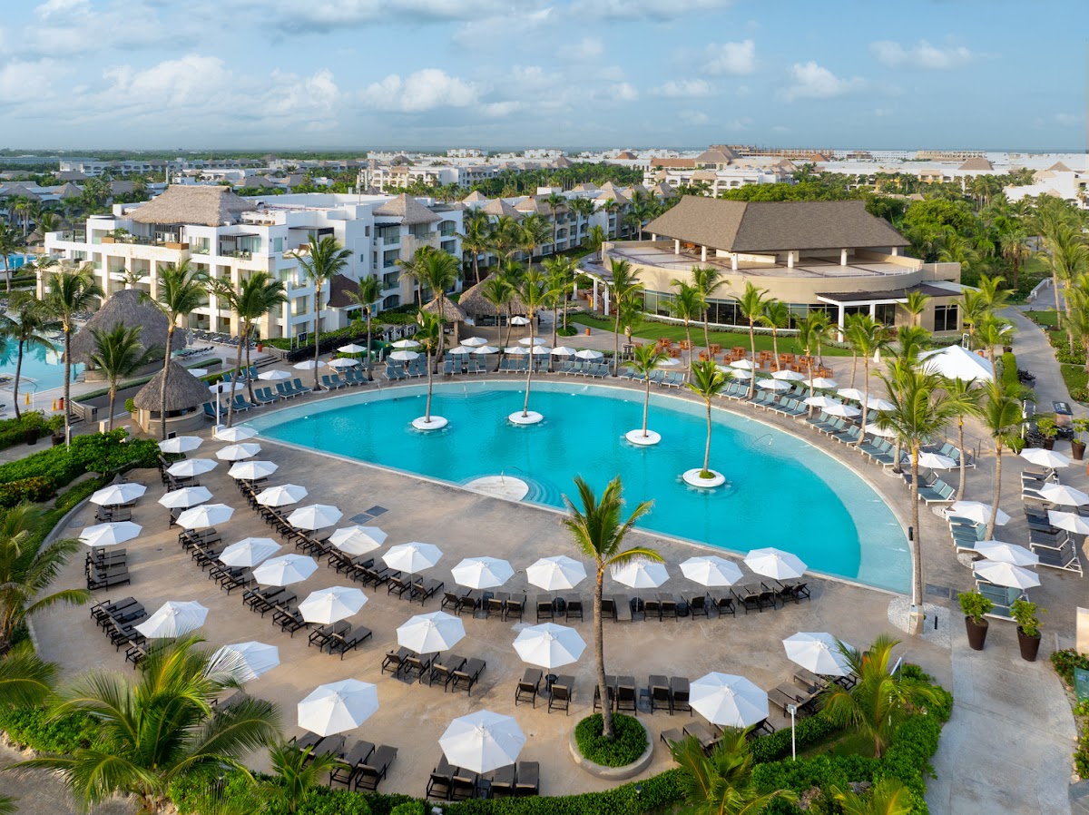 Aerial view of a large turquoise pool surrounded by white umbrellas and lounge chairs, with white resort buildings and palm trees beyond.