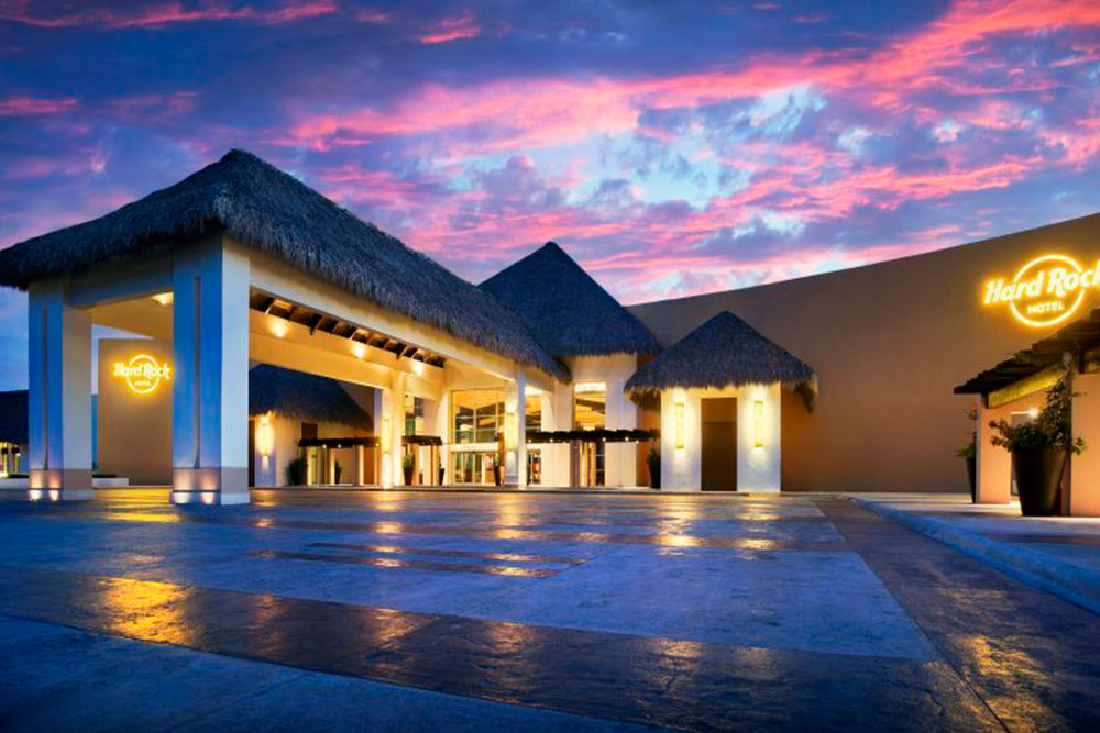 Hard Rock Hotel & Casino Punta Cana entrance at dusk with thatched-roof structures, lit columns, and dramatic pink sky.