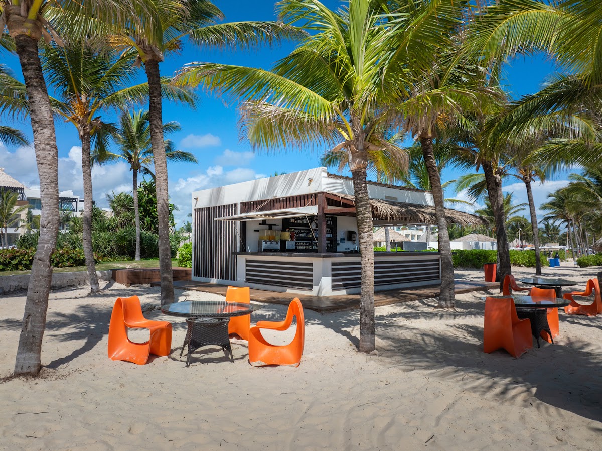 Beach bar with white and dark metal structure surrounded by palm trees and orange seating under clear blue sky at Hard Rock Hotel & Casino Punta Cana.