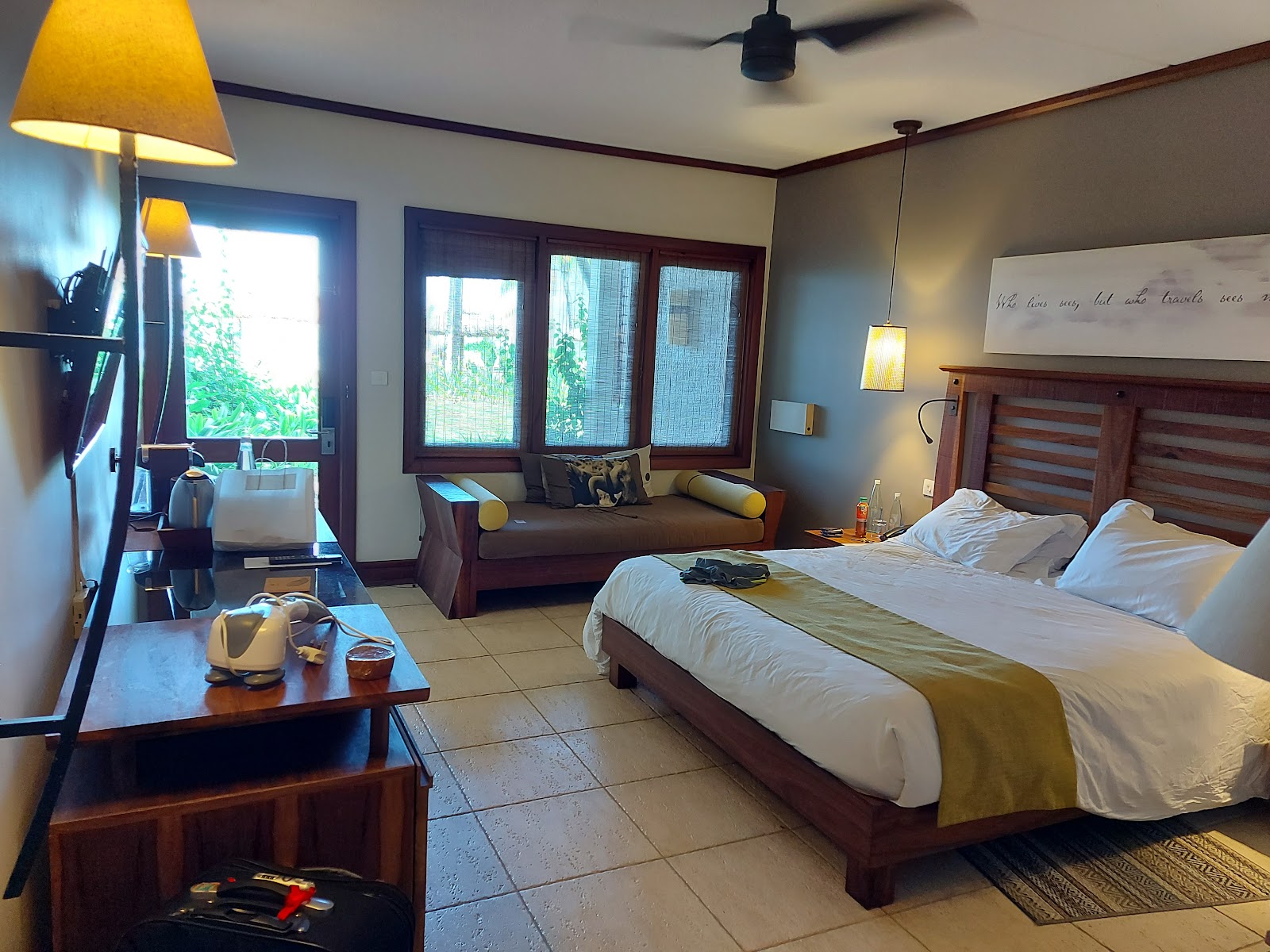 Resort suite with wooden bed, desk with TV, vanity mirror with lamps, and bathroom entrance visible in background.