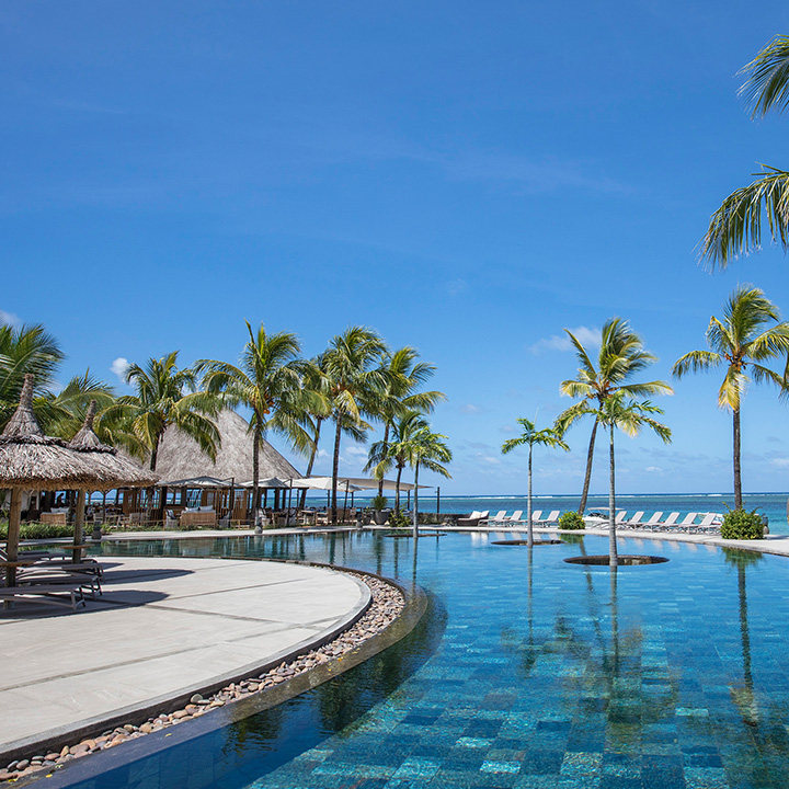 Curved pool deck with thatched-roof bar, palm trees, and ocean view at Heritage Awali Golf & Spa Resort in Mauritius.