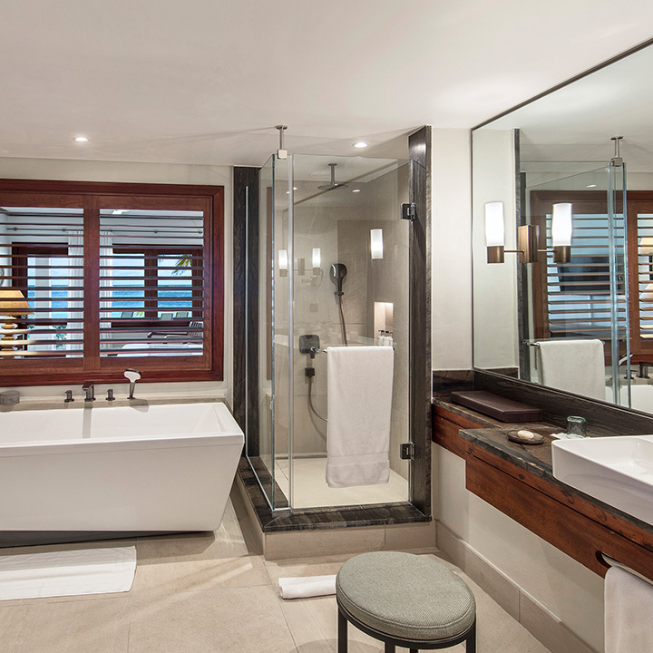 Spacious bathroom with soaking tub, walk-in shower, dual vanities with wood cabinetry, and ocean views through wooden shutters.