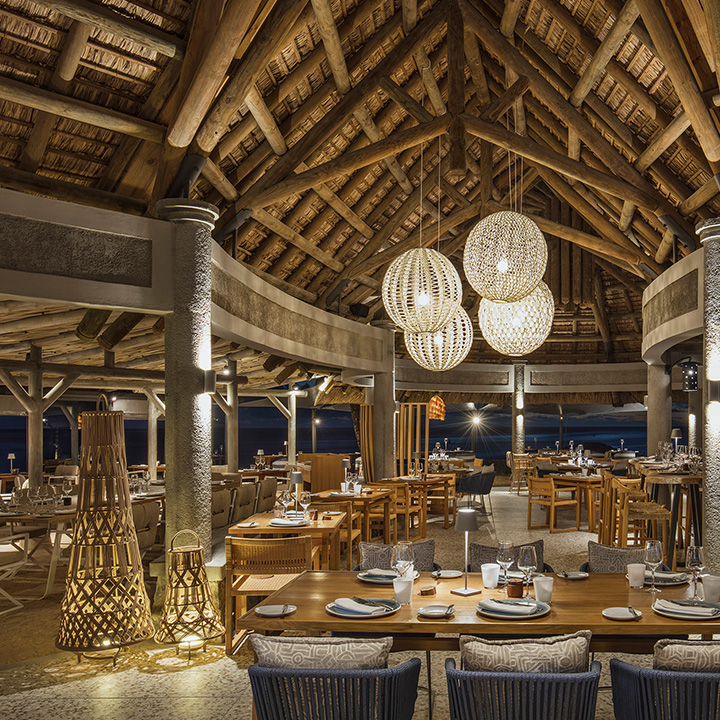 Open-air dining pavilion with thatched roof, wooden beams, pendant lights, and ocean views at Heritage Awali Golf & Spa Resort.