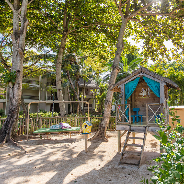 Three-story resort building with thatched roof, white walls, and wooden balconies, surrounded by palm trees and tropical plants under clear blue sky.