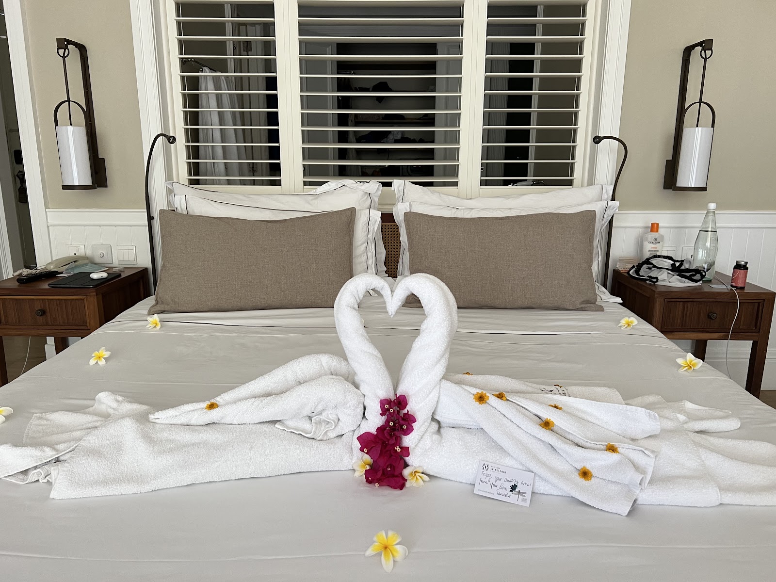 Resort room bed decorated with towel swans forming a heart shape, dark pink orchids, and yellow flowers scattered across white bedding.