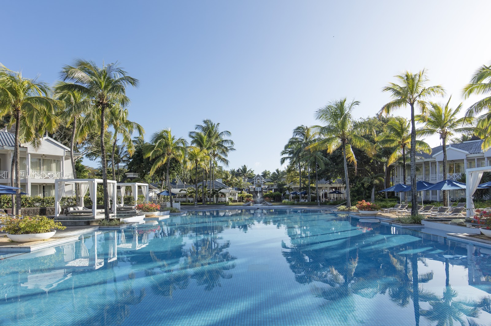 Pool with thatched umbrellas and loungers facing the ocean at Heritage Le Telfair, with palm trees and beach beyond.