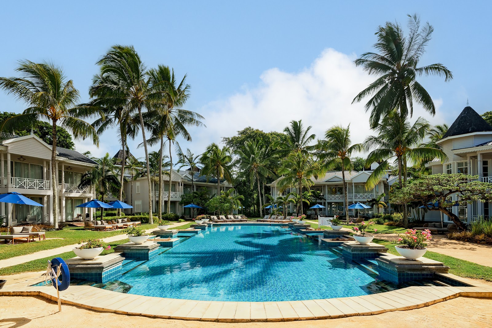 Large curved pool with blue water surrounded by colonial-style villas, palm trees, and blue umbrellas at Heritage Le Telfair Golf & Wellness Resort.