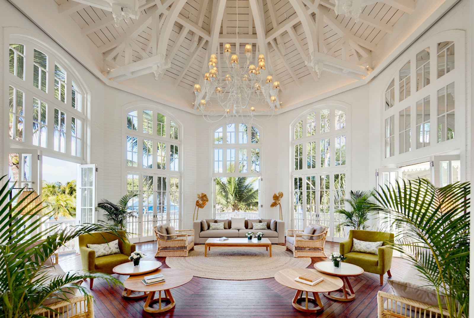 White colonial-style lounge with vaulted ceiling, crystal chandelier, palm plants, green armchairs, and French doors opening to tropical grounds at Heritage Le Telfair.