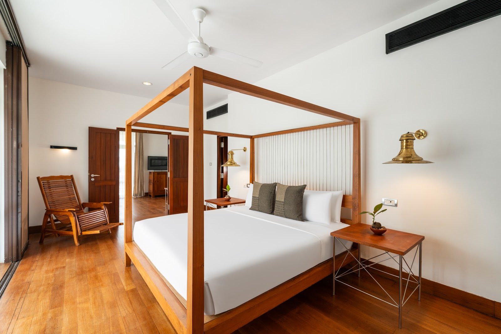 Spacious resort bedroom with wooden four-poster bed, white bedding, side table, lounge chair, and wooden floors throughout.