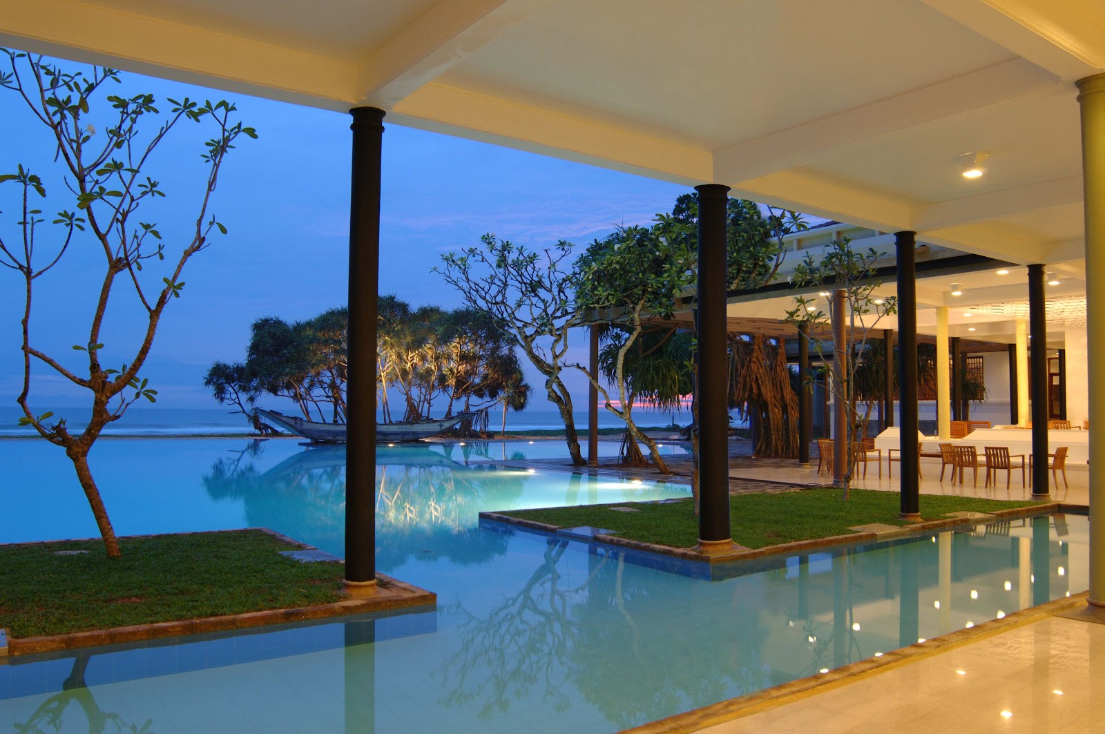 Resort pool with blue tiled loungers, palm trees, and ocean view beyond the glass barrier under clear blue sky.