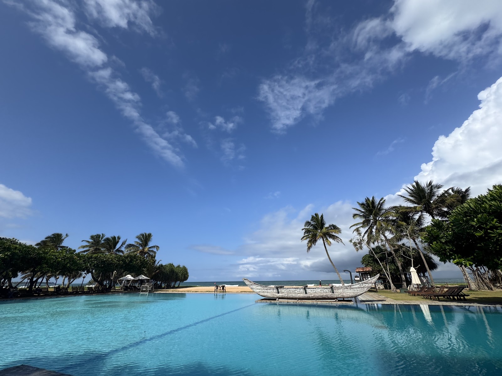 Wooden dining tables and chairs beneath palm trees overlook a turquoise lagoon pool and sandy beach at Heritance Ahungalla, with the ocean beyond.