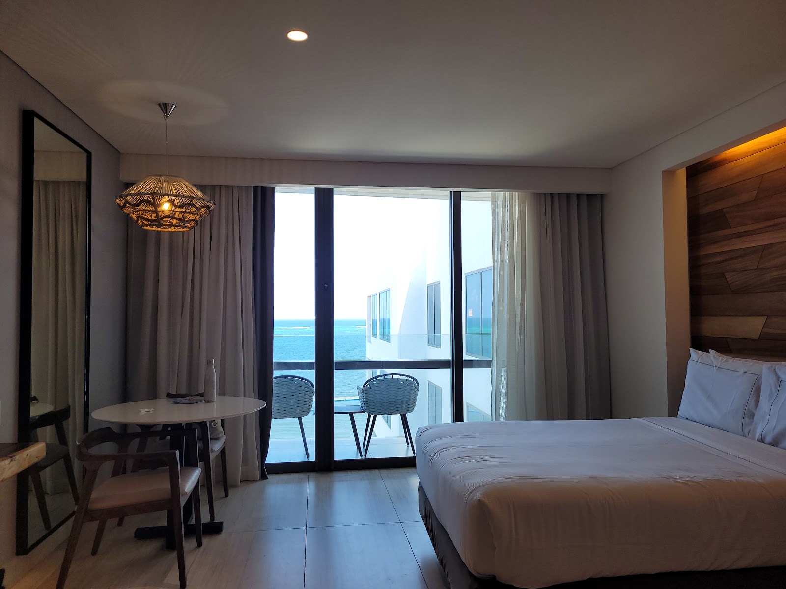 Resort guest room with two double beds, wood-paneled accent wall, pendant lighting, and ocean view through glass doors.