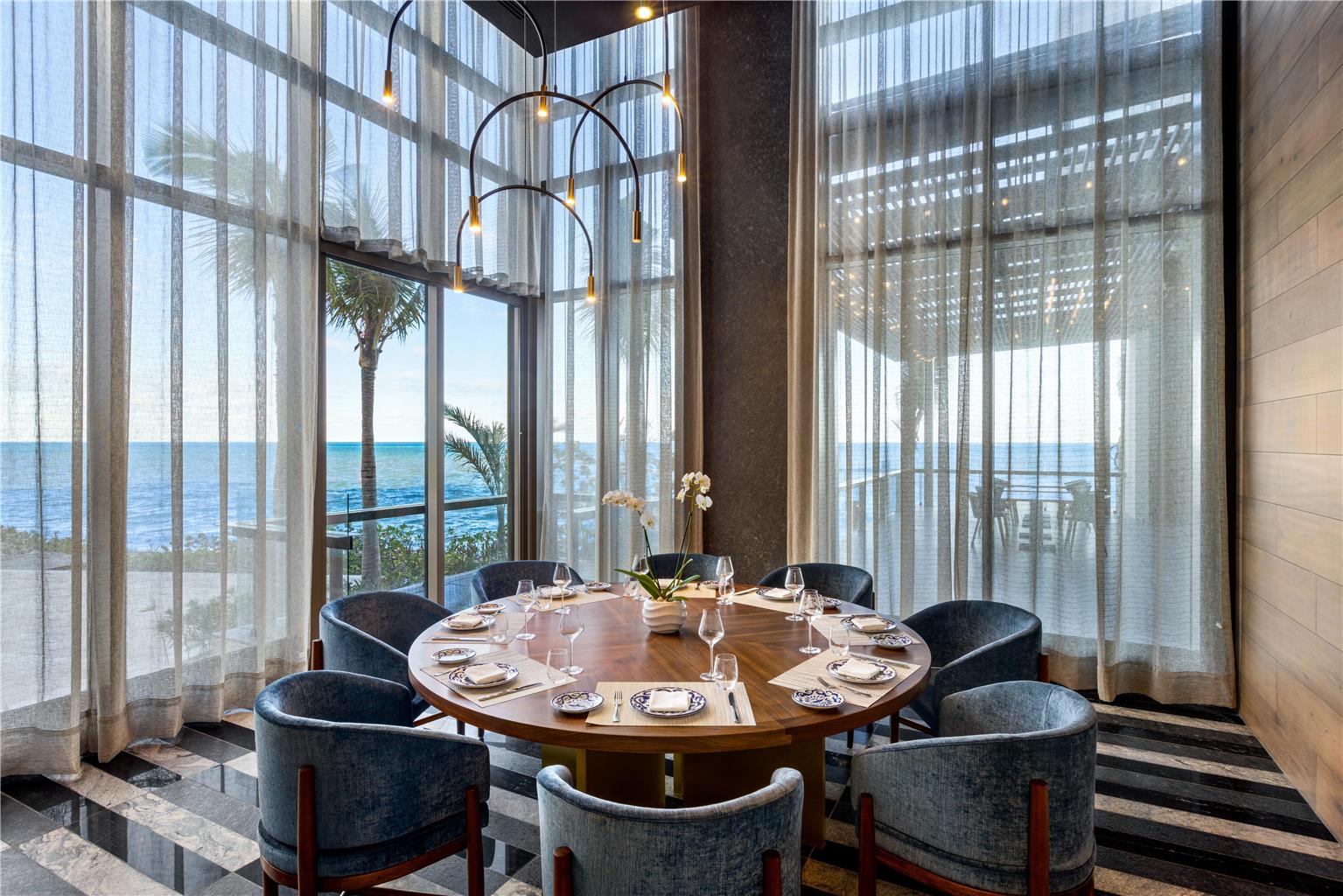 Elegant dining room with round wood table, gray upholstered chairs, and ocean views through floor-to-ceiling windows at Hilton Cancun.