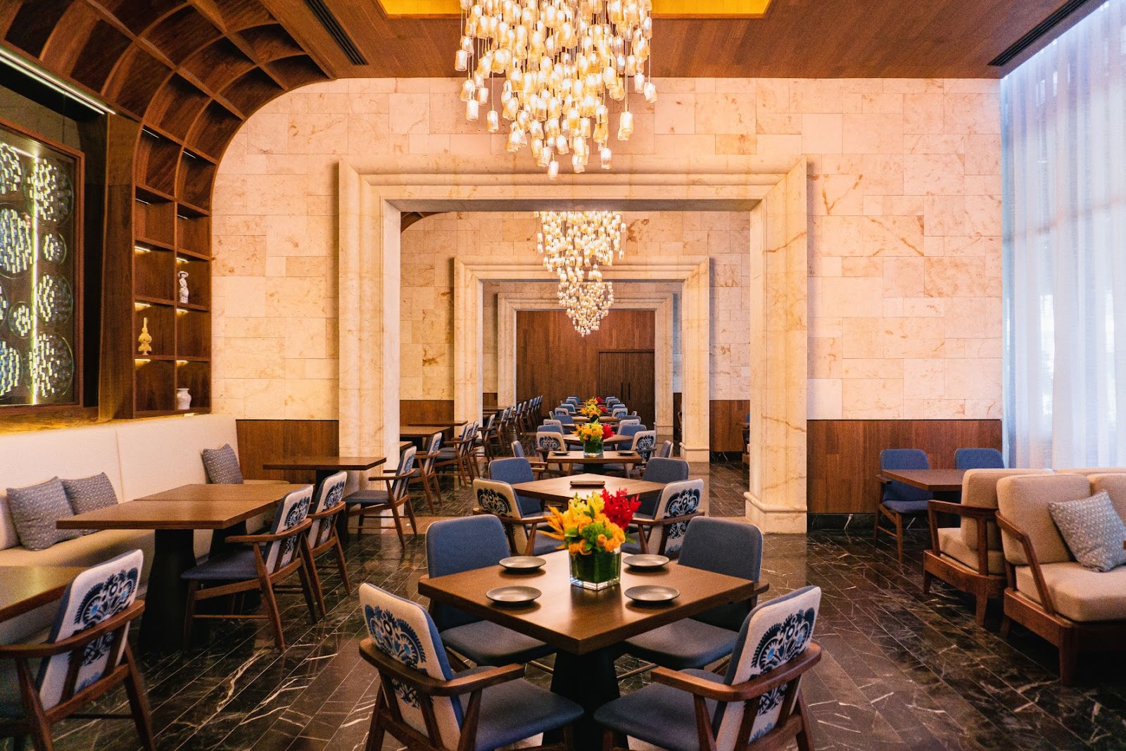 Elegant dining room with marble walls, wooden ceiling beams, crystal chandeliers, and tables set with fresh flowers and place settings.
