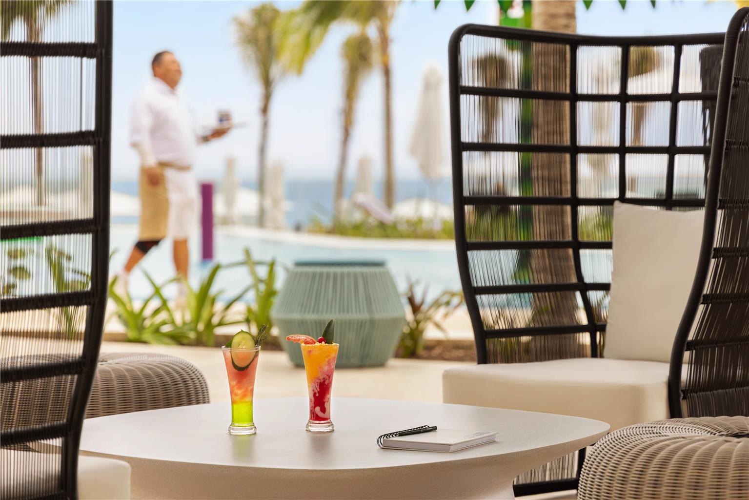 Colorful cocktails on a table surrounded by black wicker chairs, with ocean view and a man in white shirt beyond.